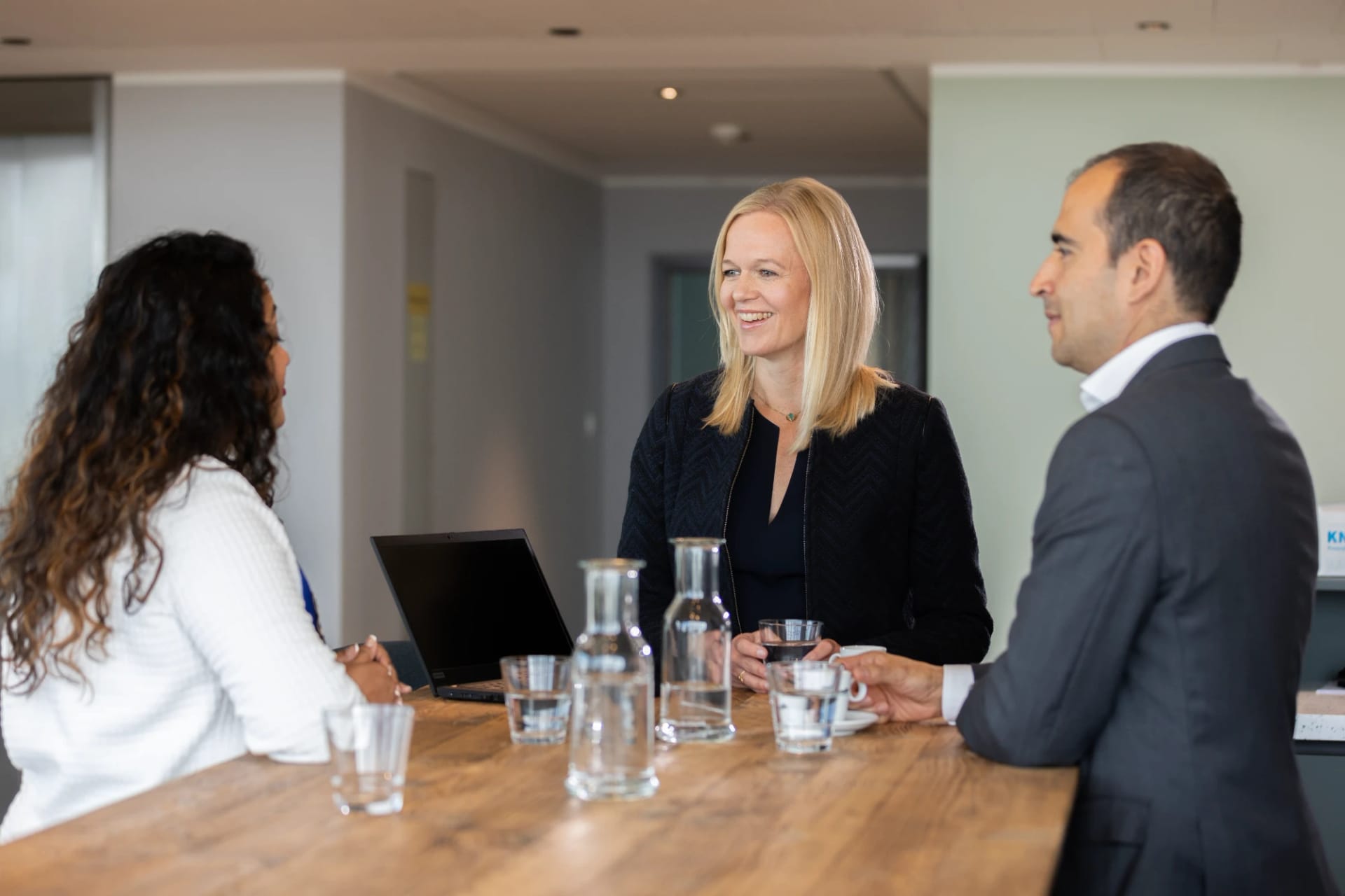 Three colleagues talk and smile at a high wooden table with a laptop.