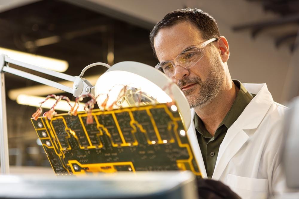 An engineer in a lab coat and safety glasses inspects a circuit board under a magnifying lamp.