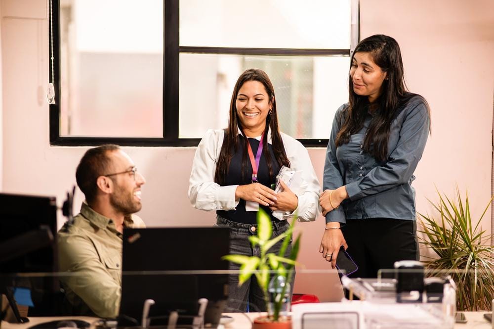 Three colleagues share a laugh at a desk in a bright office with plants.