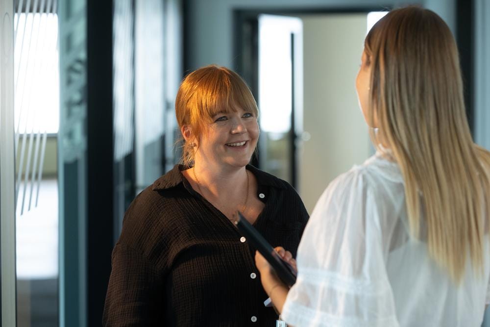 Two colleagues talk and smile in an office hallway.