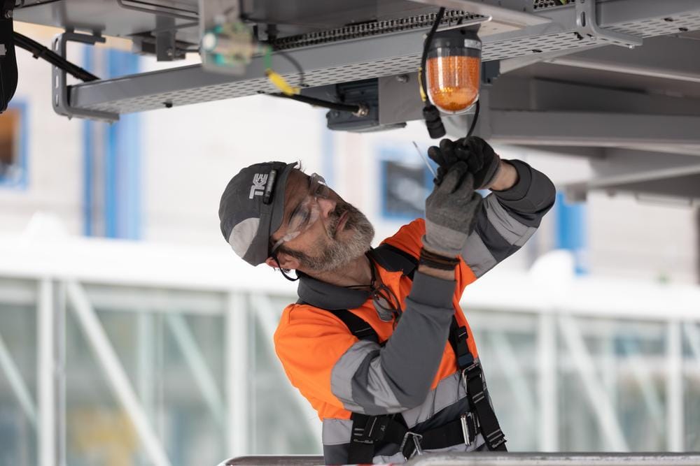 A TKE technician in safety glasses and high-visibility gear works on equipment overhead.