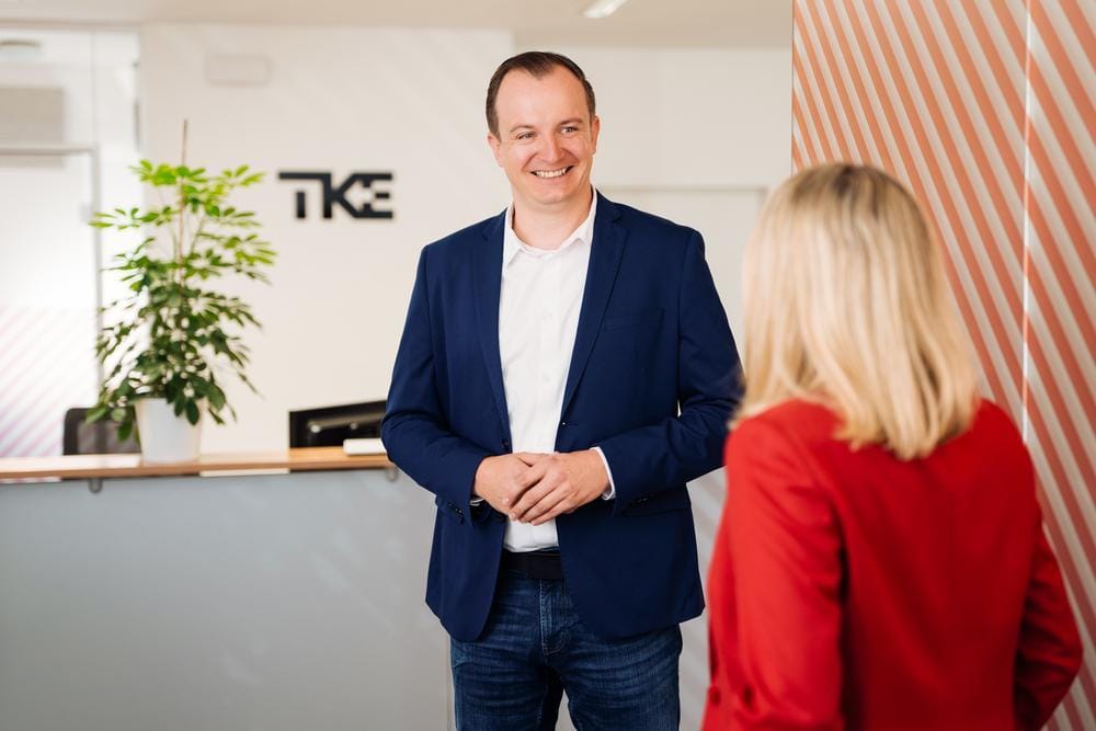 A smiling man in a navy blazer talks with a colleague in a TKE reception area.