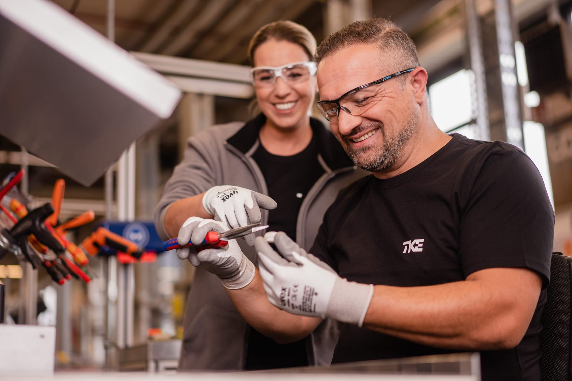 Two smiling colleagues in safety glasses and work gloves handle a tool together at a workbench.
