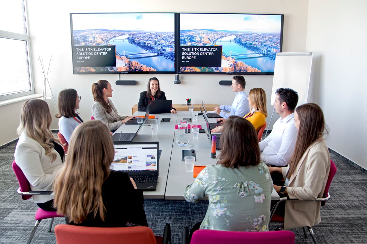 A team meets around a conference table below two screens titled “This is TK Elevator Solution Center Europe”.