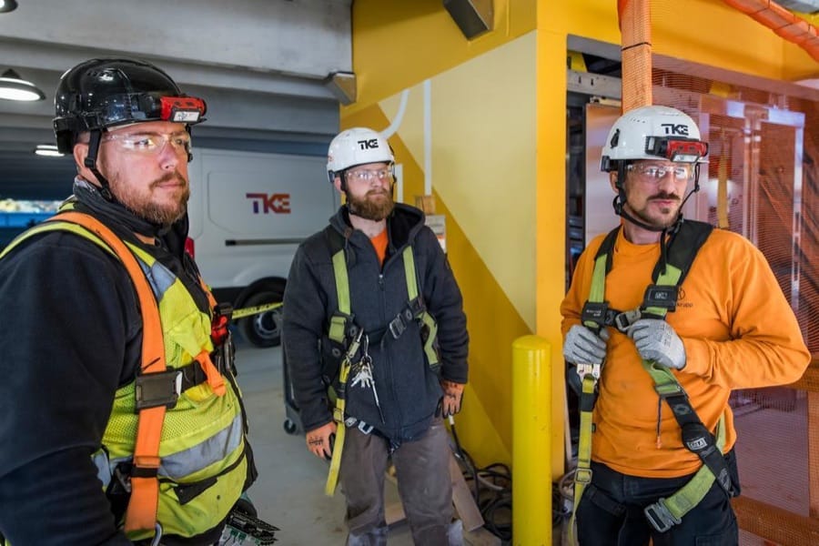 Three TKE technicians in helmets, safety glasses and harnesses on a job site.